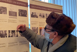 Person wearing a face mask and fur hat pointing at a display titled Daugavpils Ebreju Draudze un Kopiena