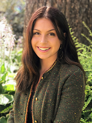 Smiling person with long dark hair wearing a textured blazer standing in a garden