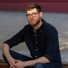 Person with glasses and a beard wearing a dark button-down shirt and jeans sitting with hands clasped